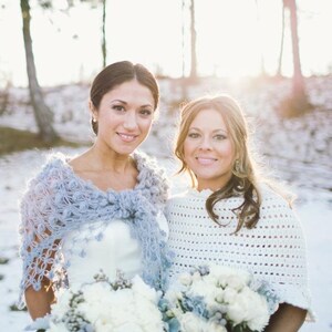 May include: Two women smiling, wearing shawls and holding bouquets of white flowers. One woman wears a light blue crocheted shawl, the other a white crocheted shawl. The background is a snowy landscape with trees.