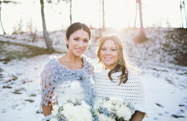 May include: Two women smiling, wearing shawls and holding bouquets of white flowers. One woman wears a light blue crocheted shawl, the other a white crocheted shawl. The background is a snowy landscape with trees.