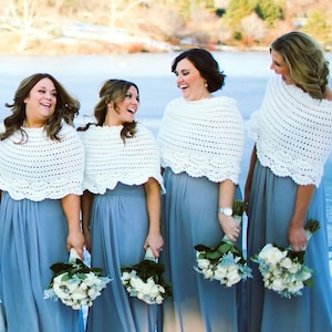 May include: Four women wearing matching outfits. Each woman is wearing a light blue dress and a white crocheted shawl. They are holding bouquets of white roses and greenery. The background shows a frozen lake and trees.