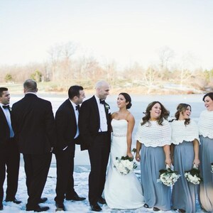 May include: A wedding party stands on a snowy landscape. The bride wears a white strapless gown, and the bridesmaids wear gray dresses and white shawls. The groomsmen wear black tuxedos. The group is smiling and laughing.