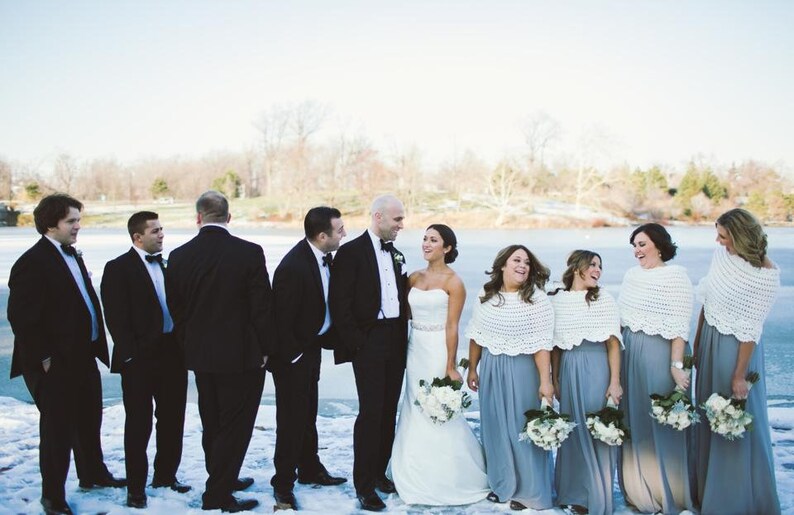 May include: A wedding party stands on a snowy landscape. The bride wears a white strapless gown, and the bridesmaids wear gray dresses and white shawls. The groomsmen wear black tuxedos. The group is smiling and laughing.