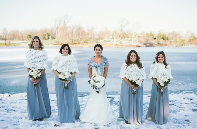 May include: Five people wearing formal attire stand in front of a frozen lake. The bridesmaids wear long, light blue dresses and white crocheted shawls. The bride wears a white dress and a blue shawl, holding a bouquet of white flowers.