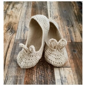May include: A pair of beige crocheted bunny slippers with a soft, fuzzy texture. The slippers have a simple design with a rounded toe and a small bunny ear detail on the top.