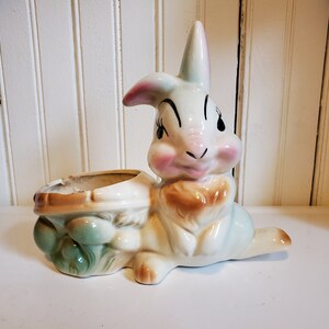 May include: A white ceramic planter shaped like a rabbit with a green and brown mushroom base. The rabbit has pink cheeks and black eyes.