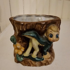 May include: A ceramic planter shaped like a tree stump with a small elf figurine peeking out from behind the stump. The elf is wearing a blue and green outfit and has large blue eyes.