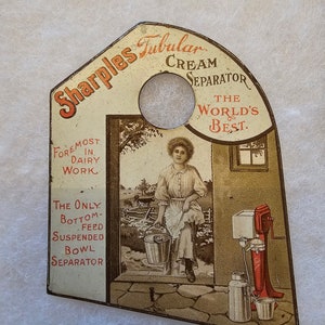 May include: A vintage tin advertising Sharples Tubular Cream Separator. The ad features a woman in a white dress and apron holding a bucket, standing in front of a barn. The text on the ad reads "Sharples Tubular Cream Separator", "The World's Best", "Foremost in Dairy Work", and "The Only Bottom-Feed Suspended Bowl Separator".