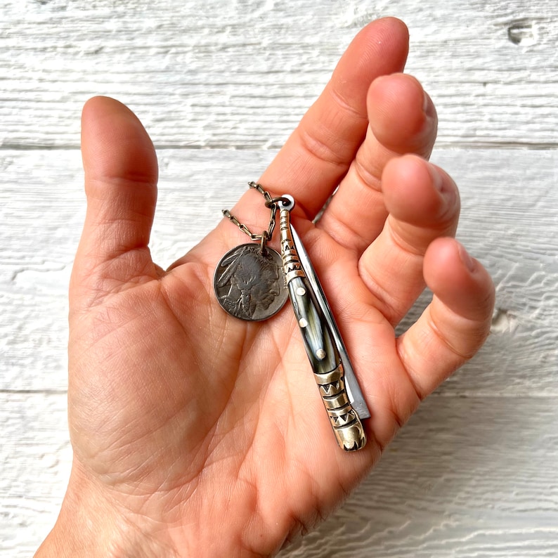May include: A silver chain necklace with a silver coin pendant featuring an image of a Native American and a silver and black pocket knife pendant.