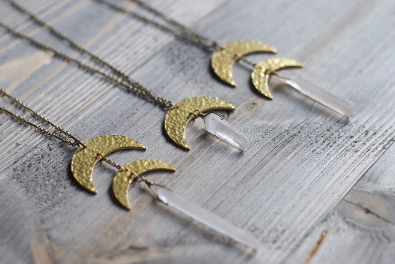 May include: Three gold necklaces with hammered crescent moon pendants and clear quartz crystal points hanging from them. The necklaces are displayed on a light wood surface.