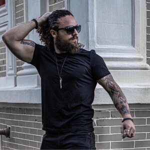 May include: A man with long curly hair and a beard is wearing a black t-shirt, black jeans, and sunglasses. He has a tattoo on his arm that says "unconditional".