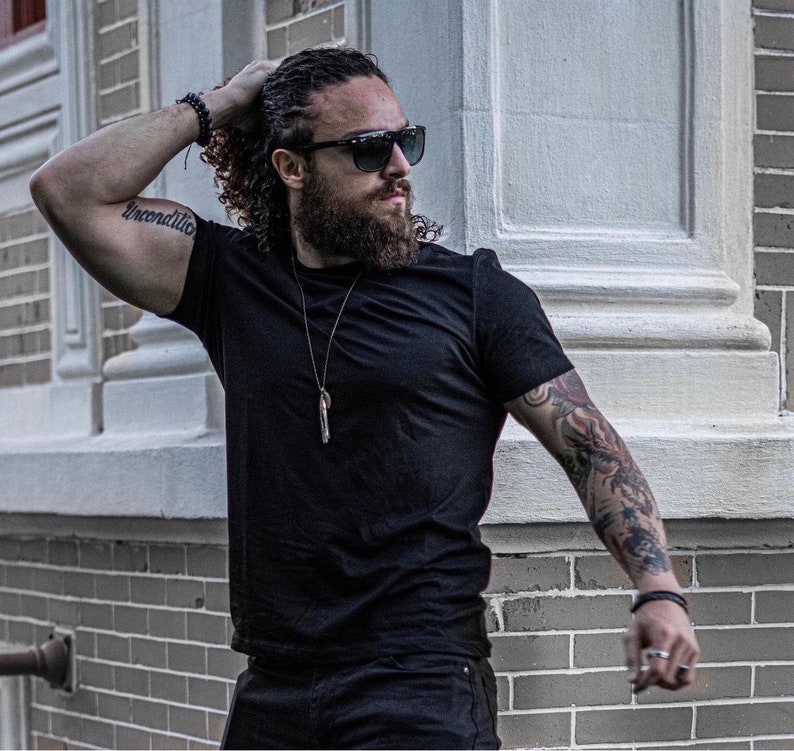 May include: A man with long curly hair and a beard is wearing a black t-shirt, black jeans, and sunglasses. He has a tattoo on his arm that says "unconditional".