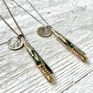 May include: Two necklaces with gold and silver chains, each featuring a vintage coin pendant and a decorative knife pendant. The knife pendants have a black and white pattern and gold accents.