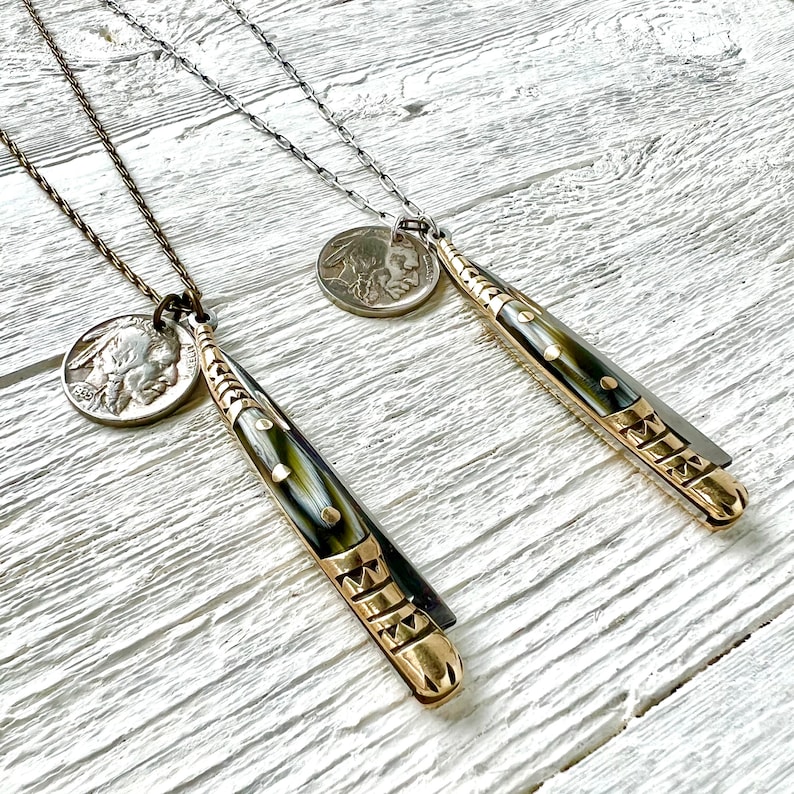 May include: Two necklaces with gold and silver chains, each featuring a vintage coin pendant and a decorative knife pendant. The knife pendants have a black and white pattern and gold accents.