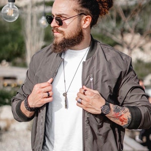 May include: A man wearing a dark gray bomber jacket with a white t-shirt underneath. He is wearing a black necklace with a pendant and a silver watch on his left wrist. He has a beard and is wearing sunglasses.