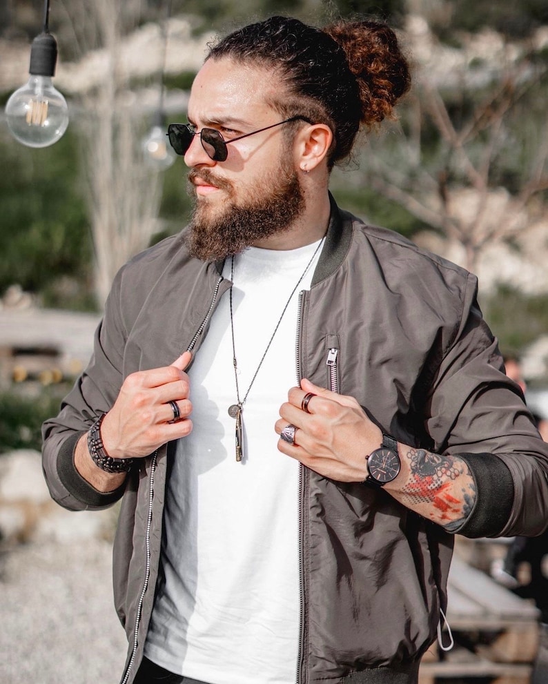 May include: A man wearing a dark gray bomber jacket with a white t-shirt underneath. He is wearing a black necklace with a pendant and a silver watch on his left wrist. He has a beard and is wearing sunglasses.