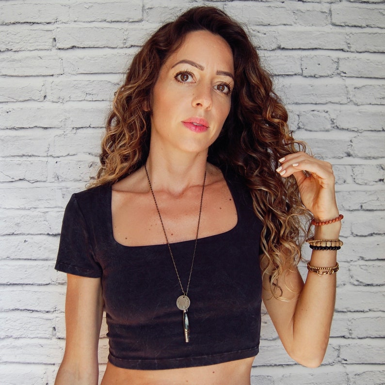 May include: A woman with long, curly brown hair wearing a black square neck crop top and a silver necklace with a round pendant. She is wearing several bracelets on her right wrist.