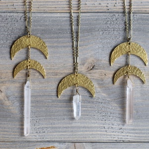 May include: Three gold necklaces with hammered crescent moon pendants and clear quartz crystal drops. The necklaces are hanging against a light wood background.