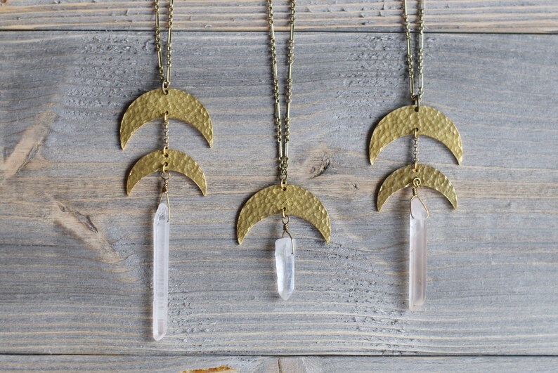 May include: Three gold necklaces with hammered crescent moon pendants and clear quartz crystal drops. The necklaces are hanging against a light wood background.