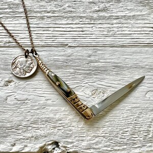 May include: A silver coin with a profile of an American Indian on it is attached to a gold chain. The chain is connected to a small, folding pocket knife with a brown and white handle. The knife is open and the blade is visible.