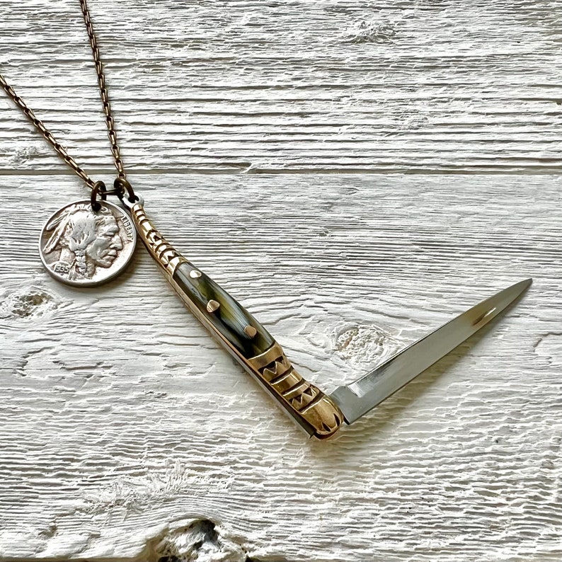 May include: A silver coin with a profile of an American Indian on it is attached to a gold chain. The chain is connected to a small, folding pocket knife with a brown and white handle. The knife is open and the blade is visible.