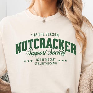 May include: Cream-colored t-shirt with the text "'TIS THE SEASON NUTCRACKER Support Society" in green. Below the text are three stars, and the phrases "NOT IN THE CAST" and "STILL IN THE CHAOS". The shirt is a casual, festive design.