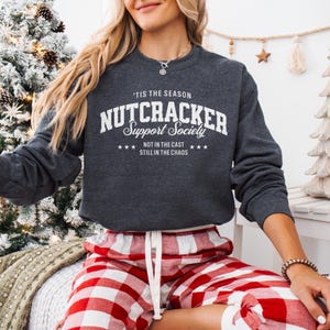 Woman wearing a dark heather gray graphic long sleeved sweatshirt with white lettering, that reads ‘’Tis the Season, Nutcracker Support Society – Not in the Cast, Still in the Chaos”. A retro collegiate-style graphic featuring bold block letters spelling ‘Nutcracker,’ script lettering for ‘Support Society,’ and smaller text below that reads ‘Not in the Cast, Still in the Chaos,’ framed by small stars.