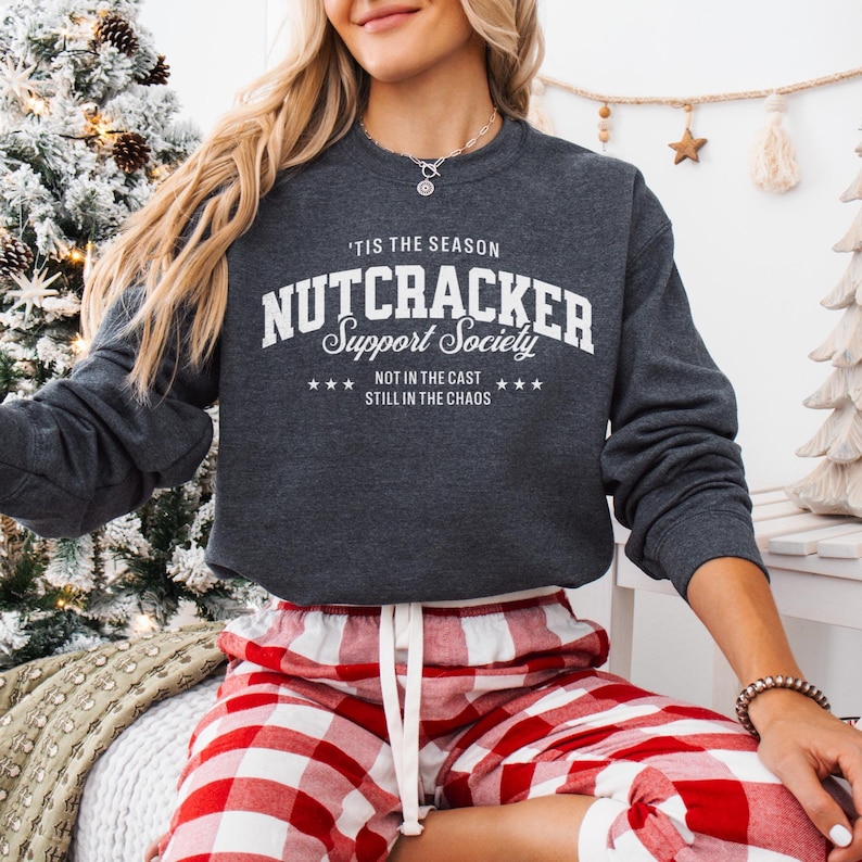 Woman wearing a dark heather gray graphic long sleeved sweatshirt with white lettering, that reads ‘’Tis the Season, Nutcracker Support Society – Not in the Cast, Still in the Chaos”. A retro collegiate-style graphic featuring bold block letters spelling ‘Nutcracker,’ script lettering for ‘Support Society,’ and smaller text below that reads ‘Not in the Cast, Still in the Chaos,’ framed by small stars.