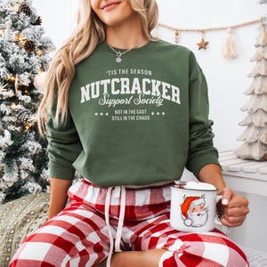 Woman wearing a medium muted green graphic long sleeved sweatshirt with white lettering, that reads ‘’Tis the Season, Nutcracker Support Society – Not in the Cast, Still in the Chaos”. A retro collegiate-style graphic featuring bold block letters spelling ‘Nutcracker,’ script lettering for ‘Support Society,’ and smaller text below that reads ‘Not in the Cast, Still in the Chaos,’ framed by small stars.