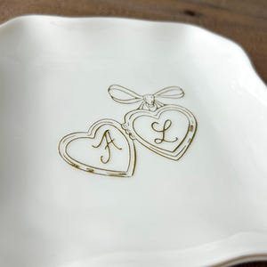 May include: A square white ceramic trinket dish with wavy edges and a gold-colored rim. The center features a gold-colored line drawing of two connected heart-shaped lockets tied with a bow, with the cursive letters A and L inside each heart.