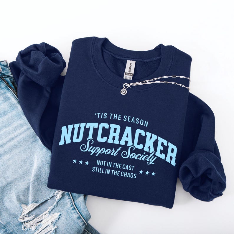 May include: A navy blue crewneck sweatshirt featuring light blue text that reads 'TIS THE SEASON NUTCRACKER SUPPORT SOCIETY NOT IN THE CAST STILL IN THE CHAOS with three stars on each side of the bottom line. A silver chain necklace rests on top.
