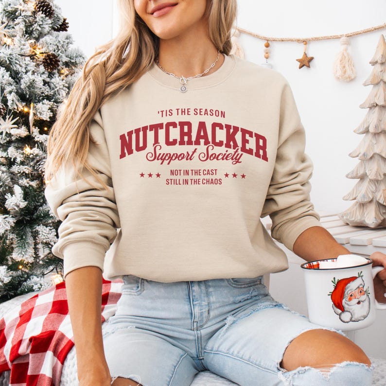 Woman wearing a beige graphic long sleeved sweatshirt with red lettering, that reads ‘’Tis the Season, Nutcracker Support Society – Not in the Cast, Still in the Chaos”. A retro collegiate-style graphic featuring bold block letters spelling ‘Nutcracker,’ script lettering for ‘Support Society,’ and smaller text below that reads ‘Not in the Cast, Still in the Chaos,’ framed by small stars.