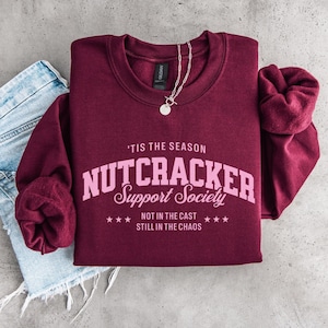 May include: A maroon long-sleeve sweatshirt featuring light pink text that reads 'TIS THE SEASON NUTCRACKER Support Society NOT IN THE CAST STILL IN THE CHAOS with small stars. A silver chain necklace rests on the collar of the sweatshirt.