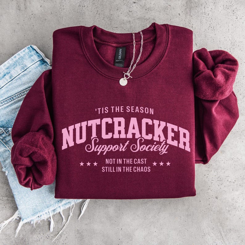 May include: A maroon long-sleeve sweatshirt featuring light pink text that reads 'TIS THE SEASON NUTCRACKER Support Society NOT IN THE CAST STILL IN THE CHAOS with small stars. A silver chain necklace rests on the collar of the sweatshirt.