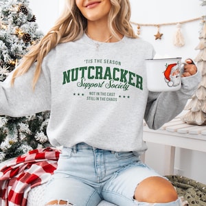 Woman wearing a light ash gray graphic long sleeved sweatshirt with green lettering, that reads ‘’Tis the Season, Nutcracker Support Society – Not in the Cast, Still in the Chaos”. A retro collegiate-style graphic featuring bold block letters spelling ‘Nutcracker,’ script lettering for ‘Support Society,’ and smaller text below that reads ‘Not in the Cast, Still in the Chaos,’ framed by small stars.