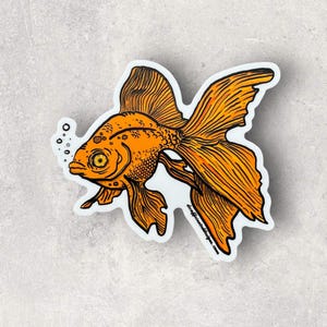 May include: A sticker featuring an orange goldfish with detailed black line art. The fish has large, flowing fins and a bubble stream. The sticker has a white border and is set against a light gray background.
