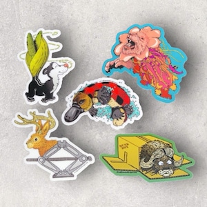 May include: Five colorful stickers featuring various animals. A platypus swimming, a deer in a car jack, a skunk with a corn cob tail, a bison in a box, and a pink creature with tentacles.