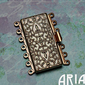 May include: A gold-toned metal jewellery clasp with an ornate design. The clasp has multiple loops for attaching jewellery chains or cords.