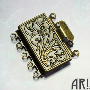May include: A rectangular antique brass jewelry clasp featuring a raised filigree scroll pattern on the top surface. The clasp includes a sliding mechanism and multiple small circular loops on both sides for attaching jewelry strands. The word ARIA is printed in the bottom right corner.