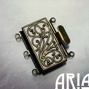 May include: A gold-toned metal jewellery clasp with a decorative filigree design. The clasp has four loops for attaching multiple strands of beads or chains.