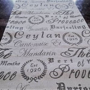 May include: Beige fabric table runner with a repeating pattern of black text and wreaths. The text includes "Ceylan", "Mandarin", "The", "Menthe", "Provence", "Aromatics", "Darjeeling", "Cantonaise", "Est 1926".