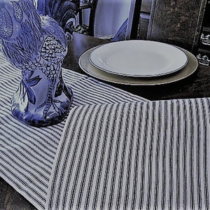 May include: A white and blue striped table runner with a blue and white ceramic rooster figurine on top. The runner is on a dark wood table with a white plate and a gold charger plate.