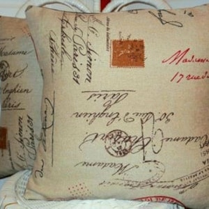May include: Two square decorative pillows with a beige background and vintage script and stamp designs in red and brown. The pillows are made of a textured fabric and are placed on a white surface.