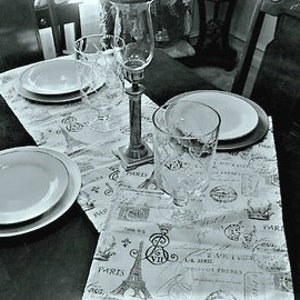 May include: A black and white table runner with a Parisian theme featuring images of the Eiffel Tower, text, and stamps. The runner is laid out on a dark wood table with white plates and glasses.