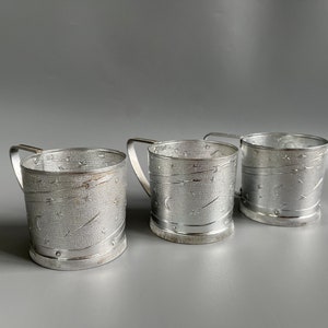May include: Three silver metal cups with a star and crescent moon design. The cups have a handle and are made of a textured metal.