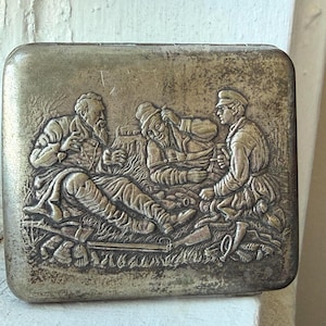 May include: A rectangular silver-toned metal case featuring a detailed embossed relief of three figures resting in a grassy field. The scene depicts individuals in period clothing with accessories like a horn and rifles, rendered in a metallic finish.