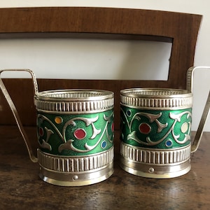 Op de afbeelding: Twee sierlijke metalen bekerhouders met groene emaille en kleurrijke accenten. De houders hebben een zilveren afwerking en decoratieve handvatten. Het ontwerp heeft bloemmotieven en is waarschijnlijk vintage.