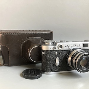 Pode incluir: Uma câmera vintage FED-2 com estojo de couro e tampa da lente. O corpo da câmera é preto e prateado, com o texto "ФЭД-2" visível. A lente tem marcações de medição. O estojo de couro marrom está ao lado da câmera.
