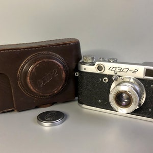 Könnte beinhalten: Eine silberne und schwarze 35-mm-Filmkamera mit einer Ledertasche. Die Kamera hat eine Objektivabdeckung und einen Sucher. Die Kamera ist mit "M3D-2" beschriftet.