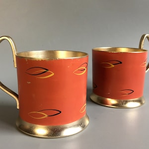 May include: Two orange metal cups with gold trim and a black and gold abstract design. The cups have a handle and are perfect for serving beverages.