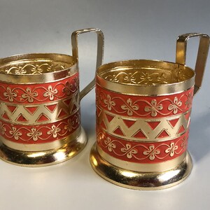 May include: Two gold-coloured metal tea glass holders with red and gold geometric designs. The holders have a handle on the side.