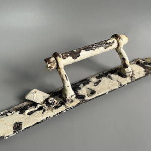 Op de afbeelding: Een antieke deurklink met een verweerde uitstraling. De klink is wit met zichtbare roest en afbladderende verf, gemonteerd op een lange, rechthoekige plaat. Het ontwerp heeft een horizontale stang ondersteund door twee gebogen palen. (ongeveer 20 cm)